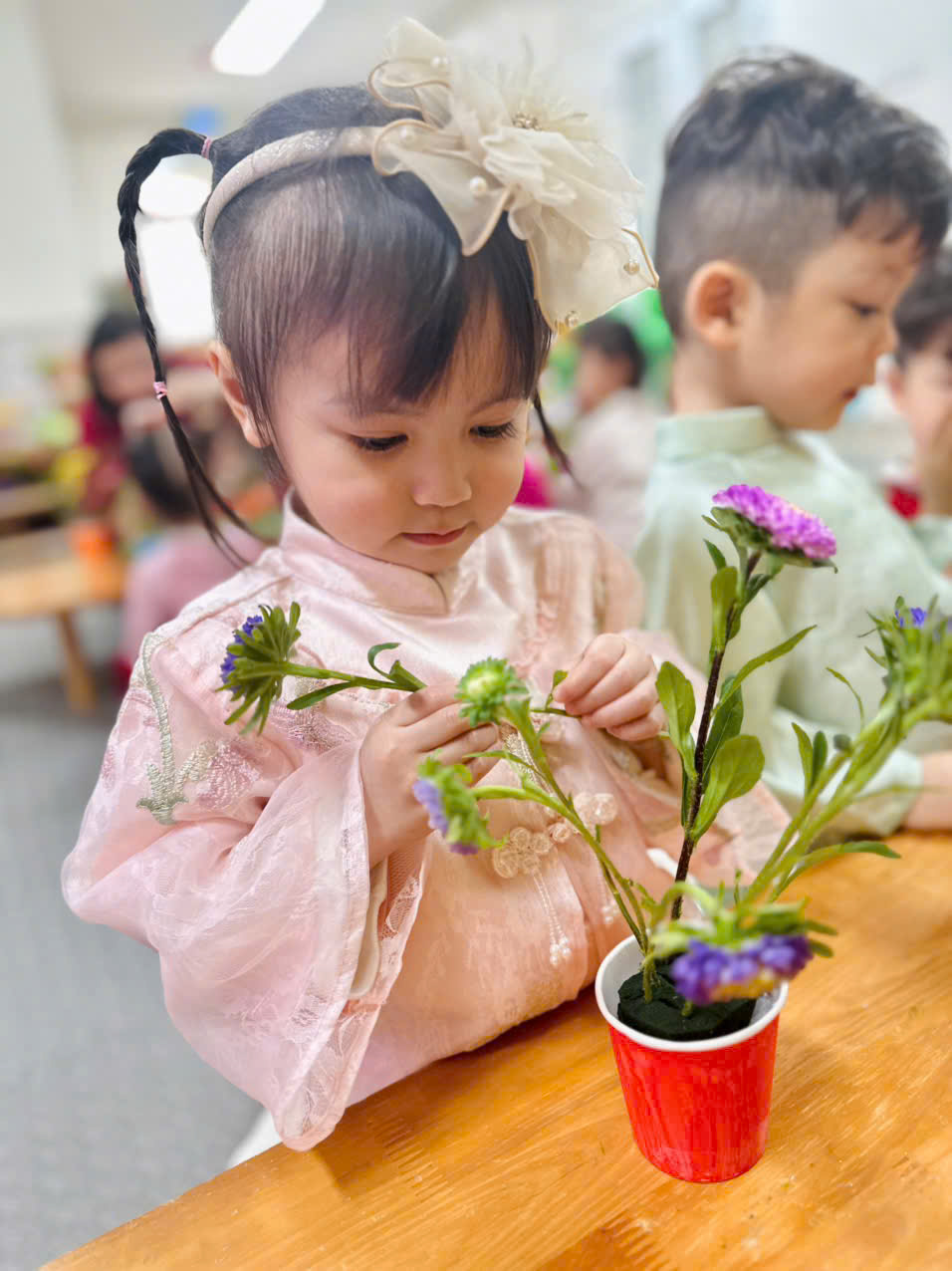 A child in a pink dress and white bow holding flowers in a red cupAI-generated content may be incorrect.
